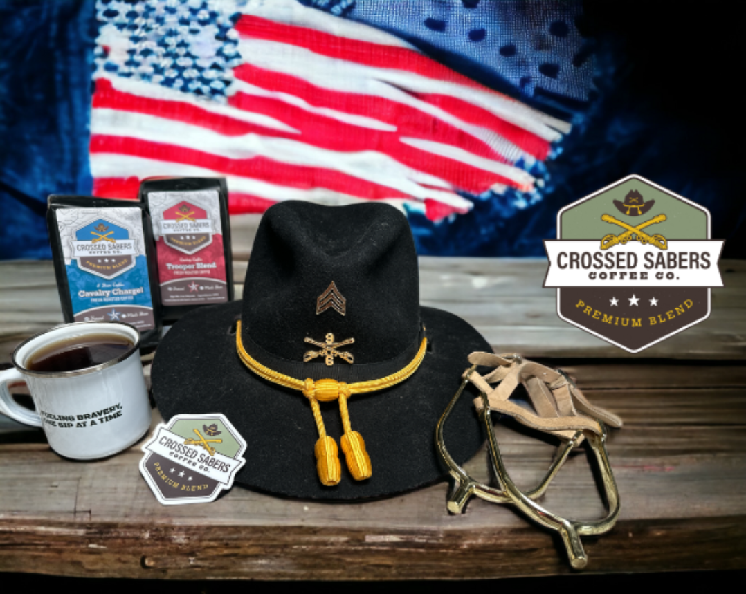 A cavalry troopers hat with a cup of coffee and two bags of Crossed Sabers coffee on a table in front of an American flag.